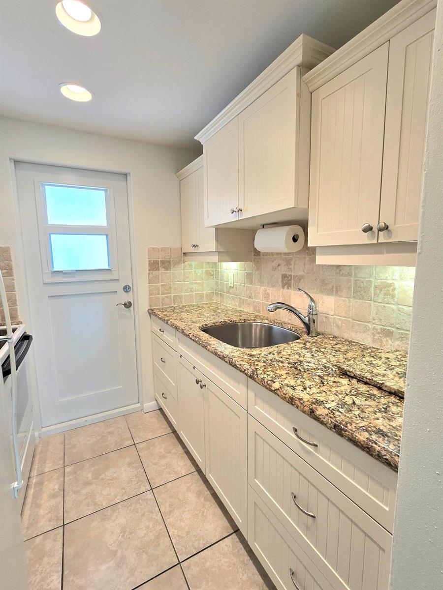 A kitchen features cream-colored cabinetry, granite countertops, a tile backsplash, and a stainless steel sink — 2501 W Golf Boulevard # 127, Pompano Beach FL 33064, POMPANO BEACH, FL