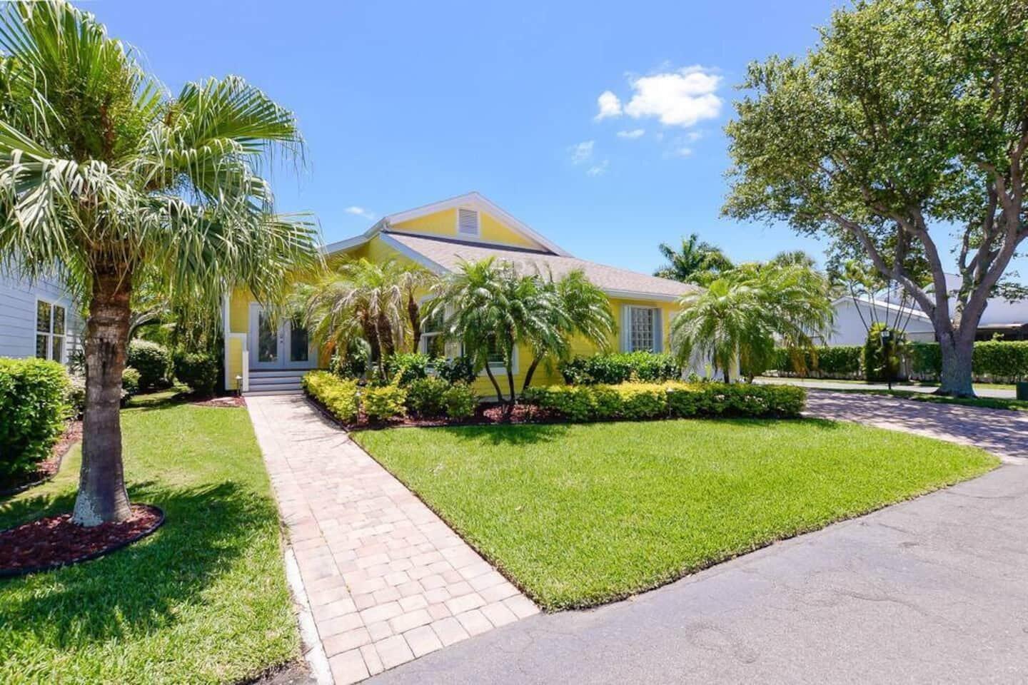 A yellow house with a brick walkway, palm trees, and a green lawn under blue skies — 4031 NE Breakwater Drive, Jensen Beach FL 34957, JENSEN BEACH, FL
