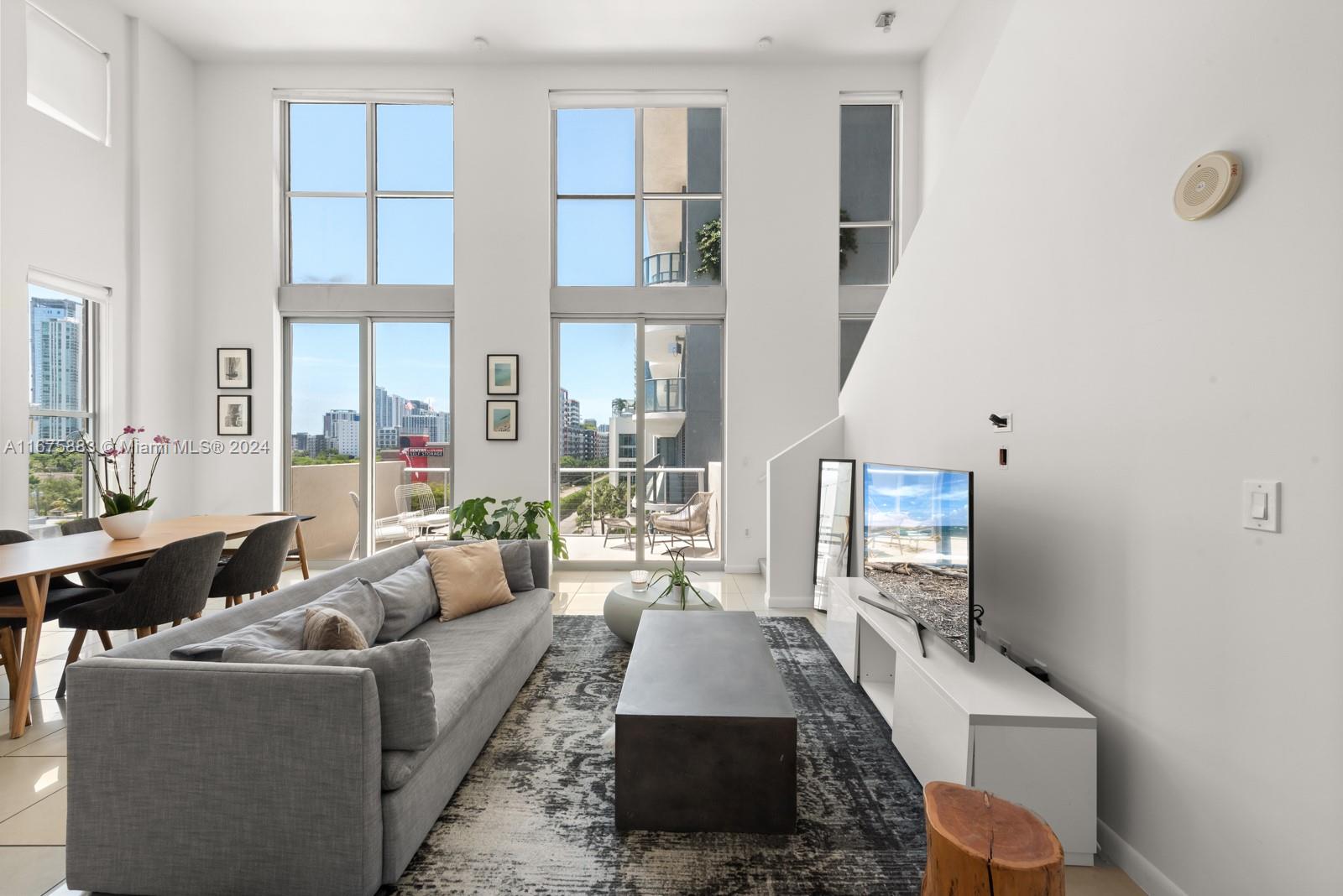 A modern living room features high ceilings, floor-to-ceiling windows, and a balcony with city views — 3449 NE 1 AVE # L61, Miami FL 33137, MIAMI, FL