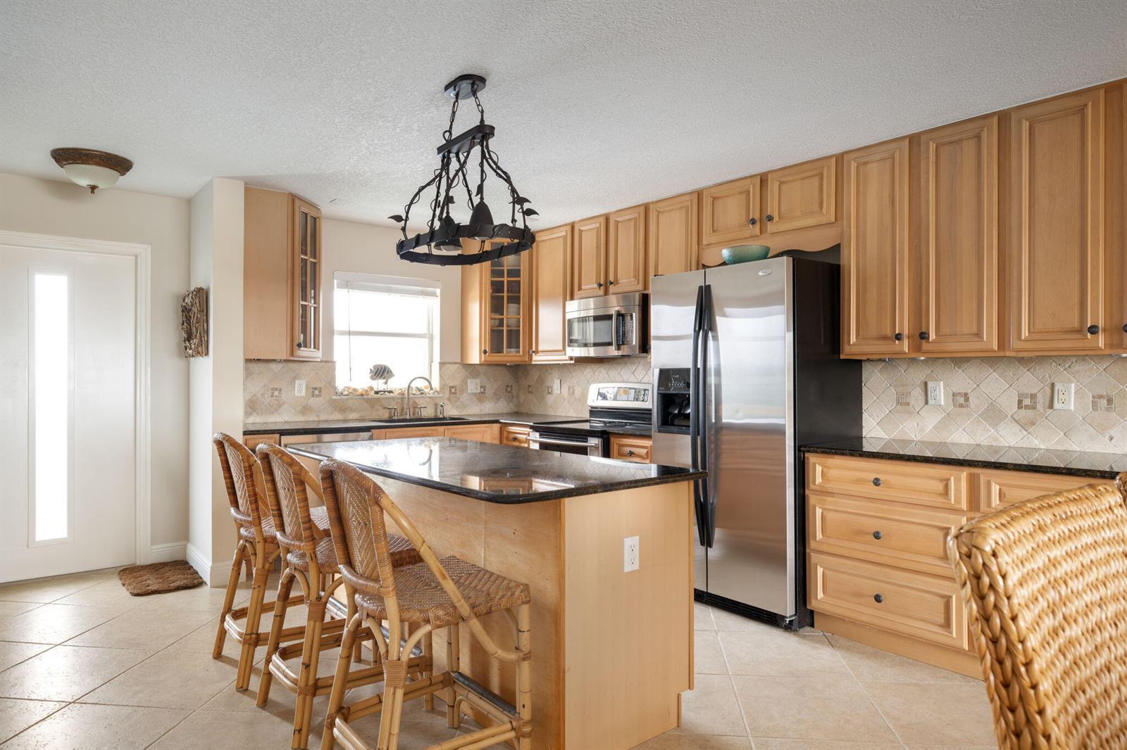 A kitchen features light wood cabinets, a granite-topped island with rattan stools, and stainless appliances — 4049 Ocean Drive # 505, Vero Beach FL 32963, VERO BEACH, FL