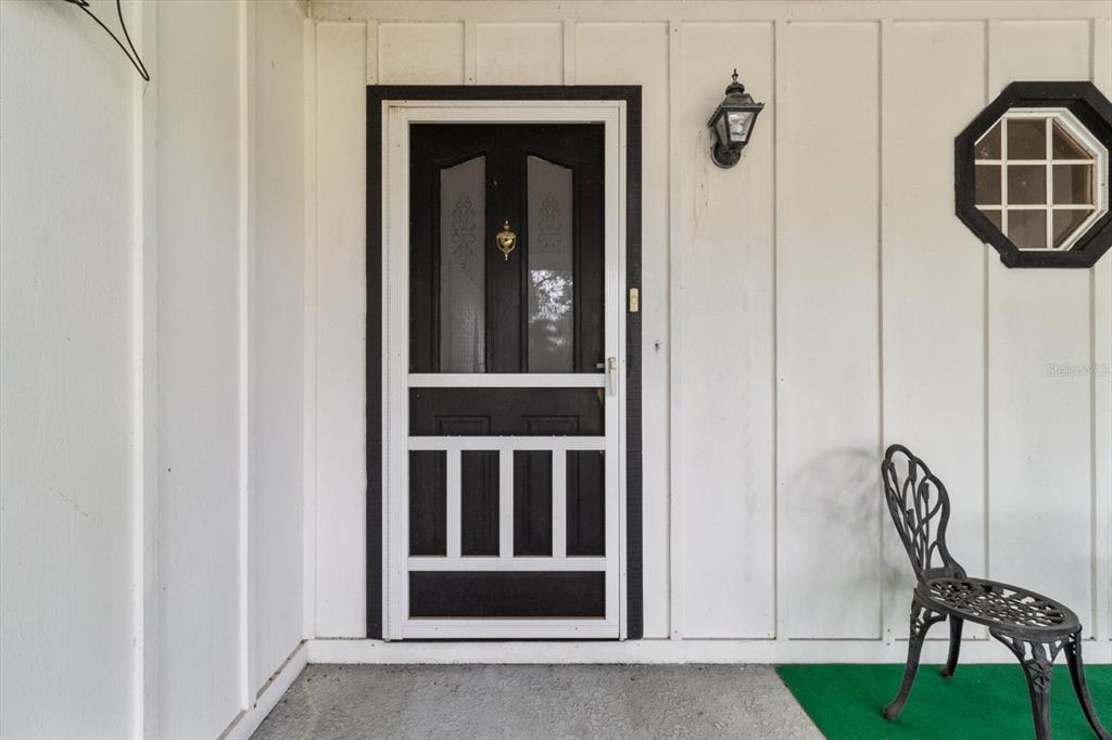 A black storm door is centered on a white exterior wall next to a metal chair — 10025 KIMBROUGH DRIVE, BROOKSVILLE, FL