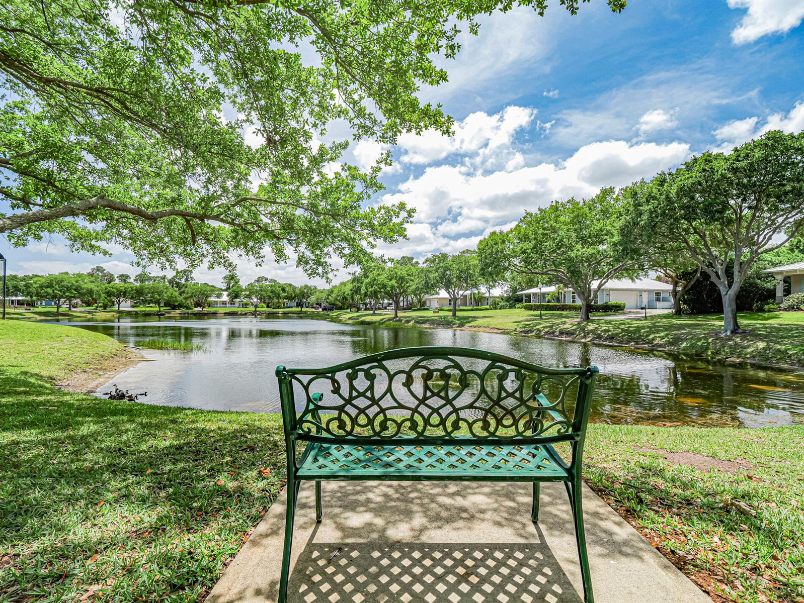 A green metal bench sits on a concrete path overlooking a pond and grassy yard — 6489 53rd Circle, Vero Beach FL 32967, VERO BEACH, FL