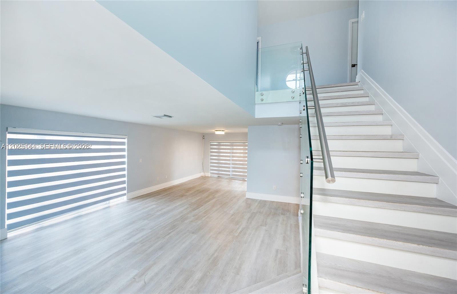 A modern living room features light wood flooring, pale blue walls, and a glass-railed staircase — 18660 NW 78th Ave # 0, Hialeah FL 33015, HIALEAH, FL