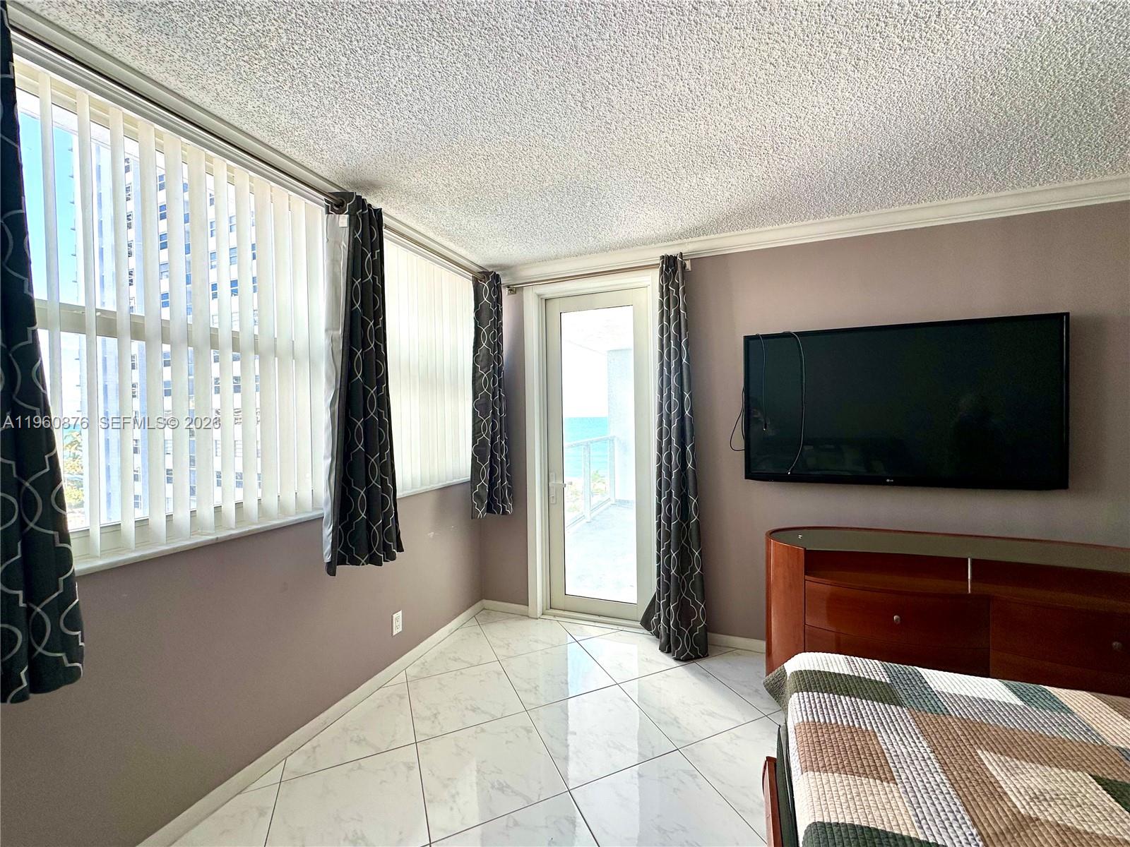 A bedroom features a wall-mounted television, tile flooring, and a glass door to a balcony — 1904 S Ocean Dr # 701, Hallandale Beach FL 33009, HALLANDALE BEACH, FL