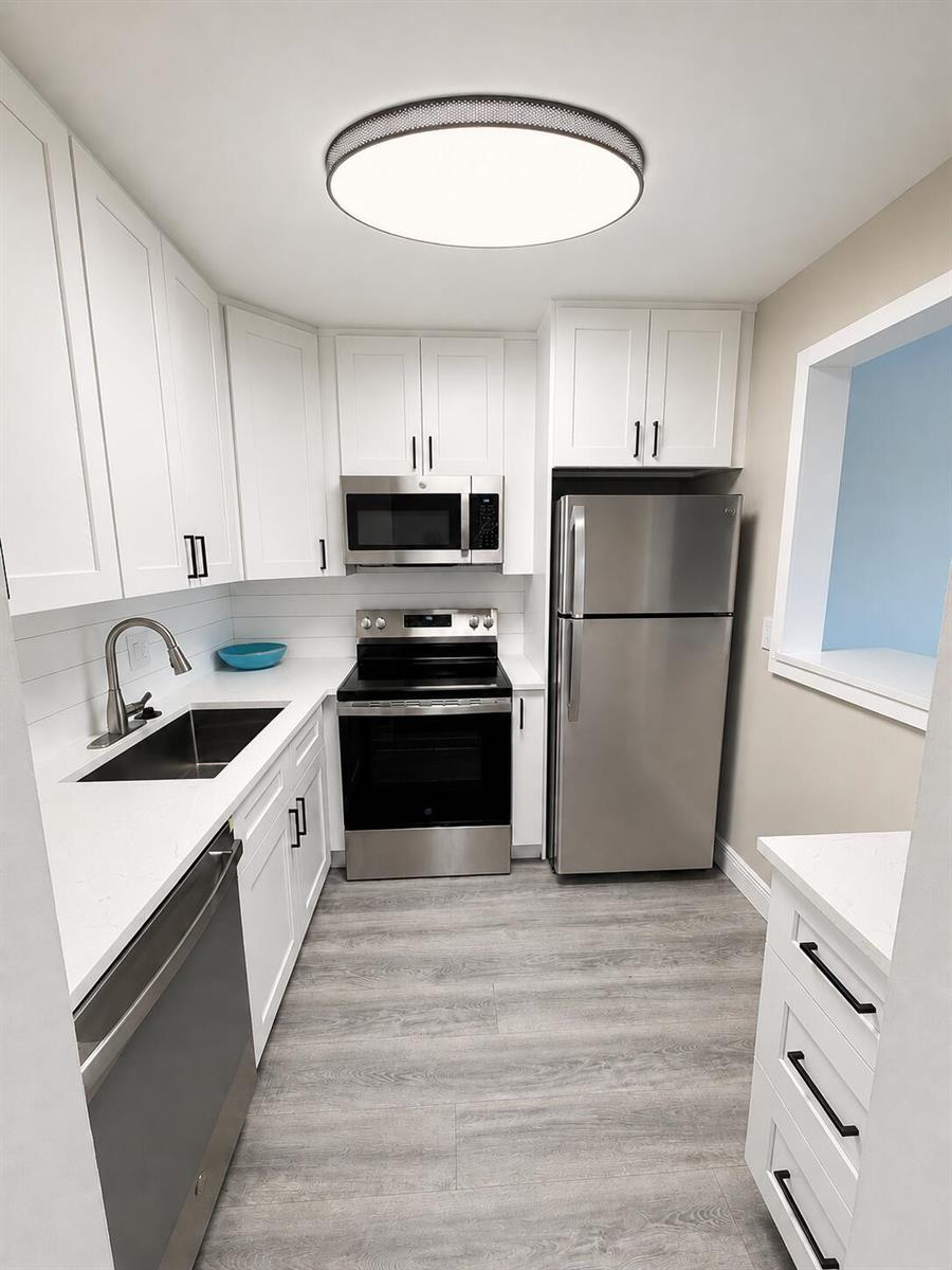 A galley kitchen features white cabinets, stainless steel appliances, quartz countertops, and gray wood flooring — 115 S South Boulevard # 2c, Boynton Beach FL 33435, BOYNTON BEACH, FL