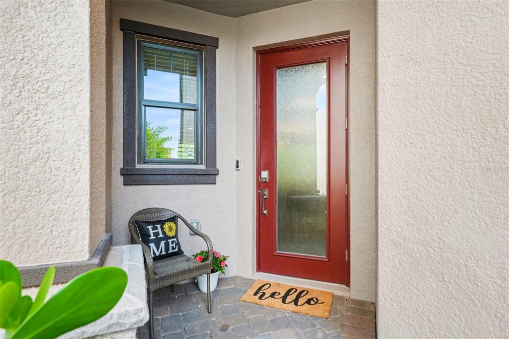 A red front door with a frosted glass panel sits beside a window and chair — 5054 SAN MARTINO DRIVE, WESLEY CHAPEL, FL