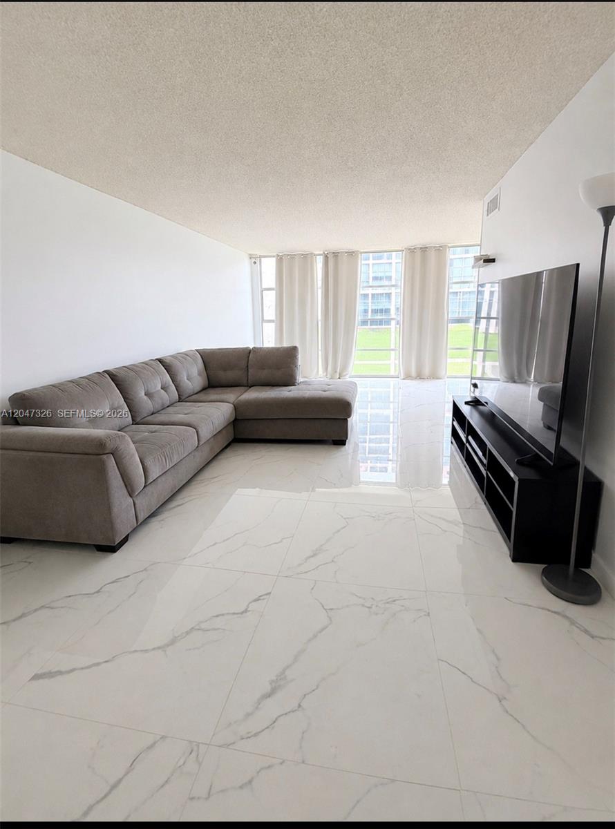 A living room features a grey sectional sofa, a black media console, and white marble-look flooring — 801 Three Islands Blvd # 107, Hallandale Beach FL 33009, HALLANDALE BEACH, FL
