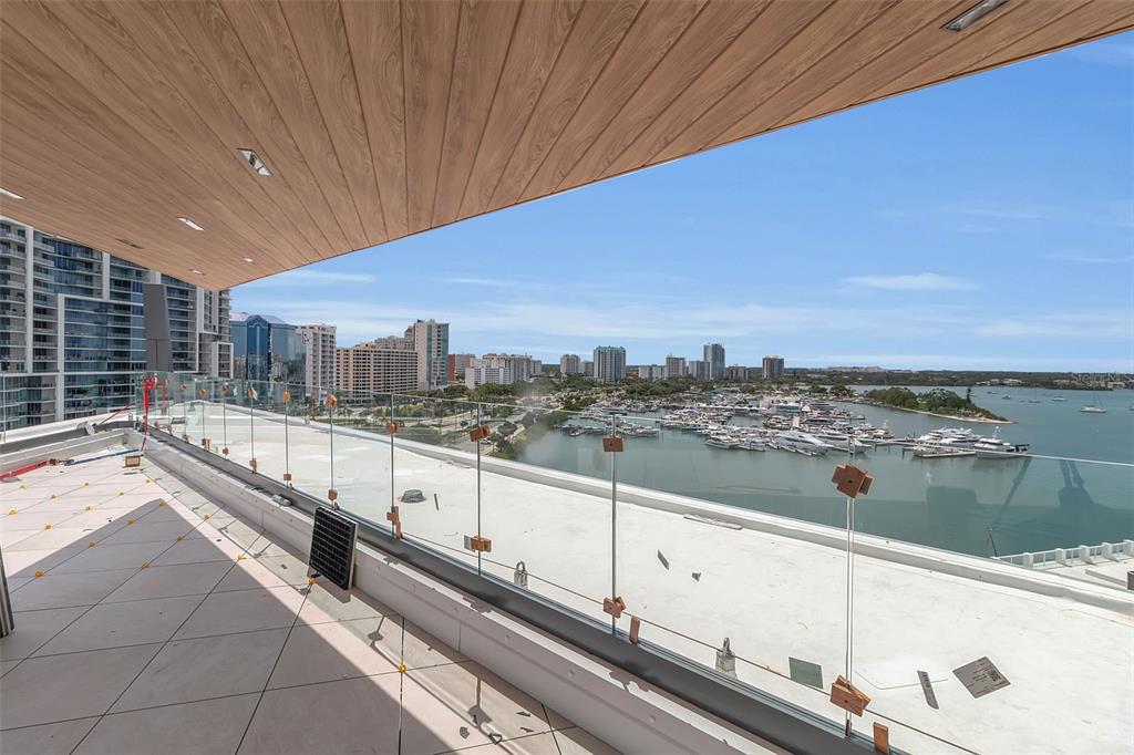 A high-rise balcony with tile flooring, a wood-paneled ceiling, and a glass railing overlooking a marina — 688 GOLDEN GATE POINT UNIT 801, SARASOTA, FL