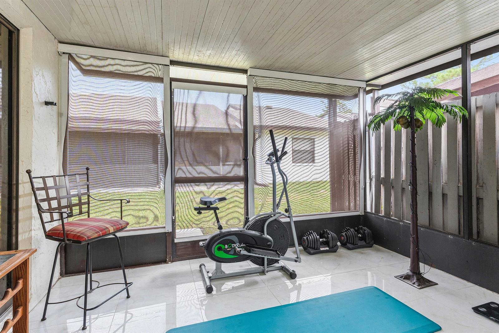 A screened-in porch with tile flooring contains an exercise bike, dumbbells, and a decorative palm tree — 1875 Abbey Road, West Palm Beach FL 33415, WEST PALM BEACH, FL