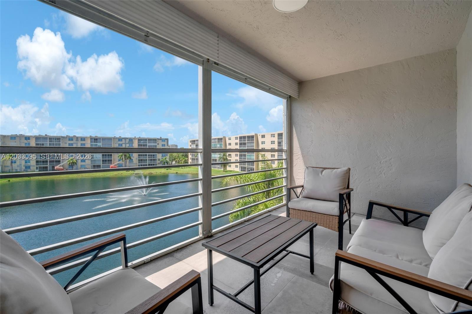 A screened balcony features patio furniture overlooking a lake with a fountain and apartment buildings — 441 SE 3rd St # 404, Dania Beach FL 33004, DANIA BEACH, FL