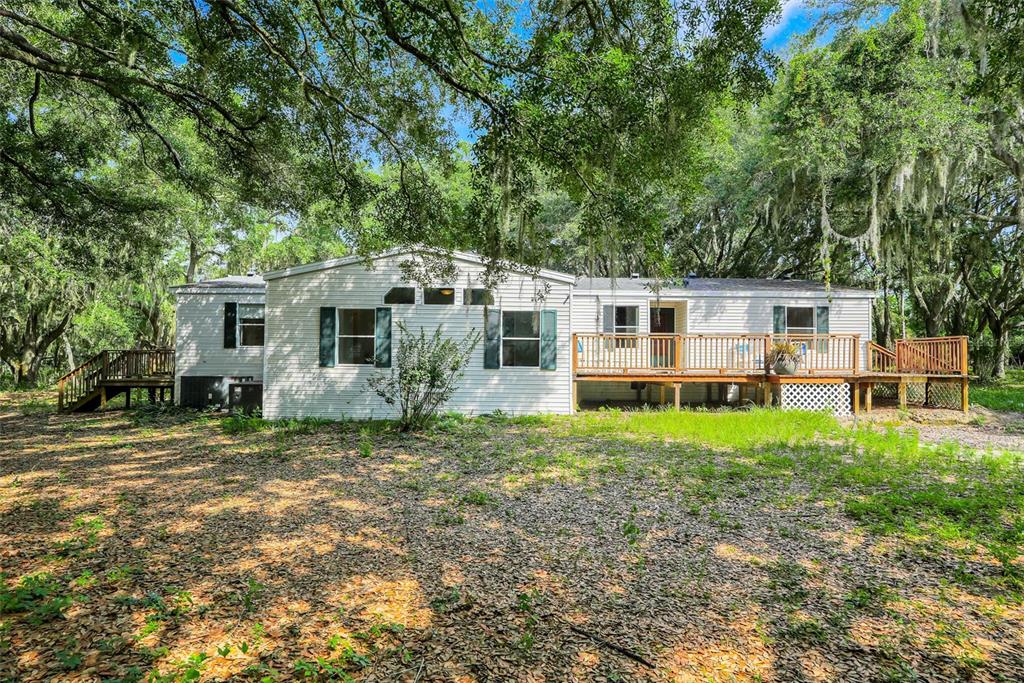 A single-story manufactured home with light siding, a wooden deck, and surrounding oak trees — 23449 THOMAS ALLEN ROAD, HOWEY IN THE HILLS, FL