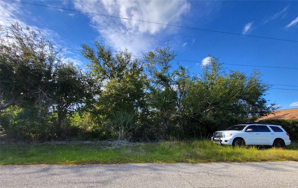 A white SUV is parked in front of a wooded, undeveloped lot under a blue sky — 9213 & 9221 GENESEE STREET, PORT CHARLOTTE, FL