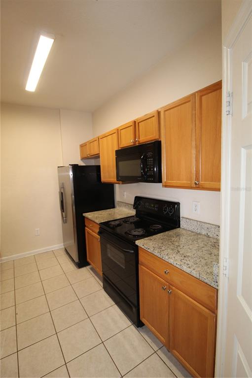 A galley kitchen features light wood cabinets, granite countertops, and black appliances on tiled flooring — 6380 CONTESSA DRIVE UNIT 208, ORLANDO, FL