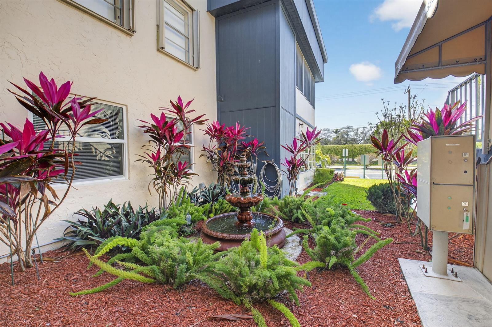 A tiered water fountain sits in a garden bed with red mulch and tropical plants — 173 Flanders D, Delray Beach FL 33484, DELRAY BEACH, FL