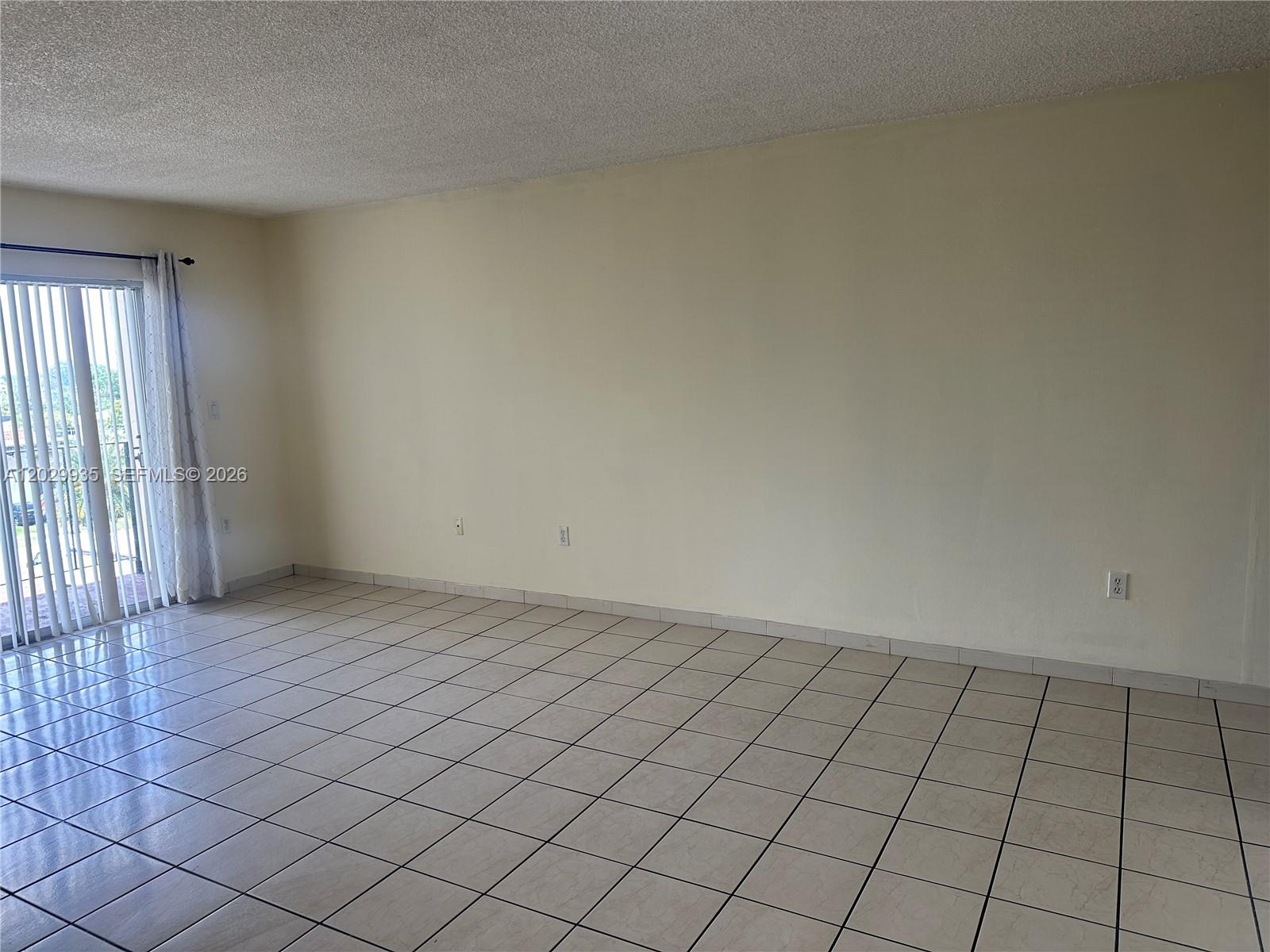 An empty room features beige tile flooring, light-colored walls, and a sliding glass balcony door — 1075 W 68th St # 309, Hialeah FL 33014, HIALEAH, FL