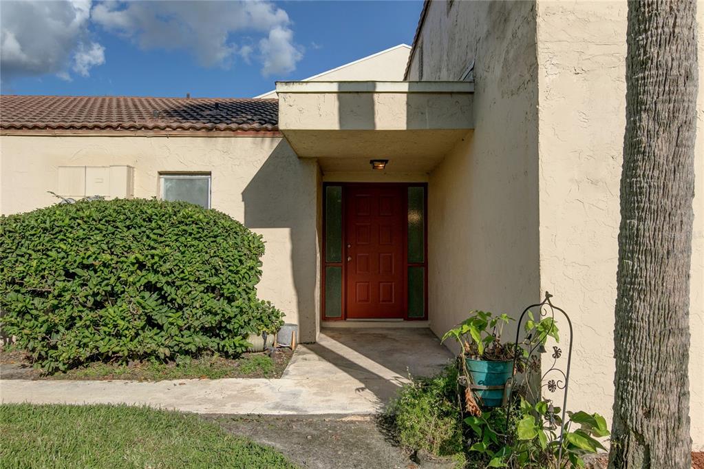 A red front door is recessed under a covered entryway next to a large bush — 602 DESOTO DRIVE, CASSELBERRY, FL
