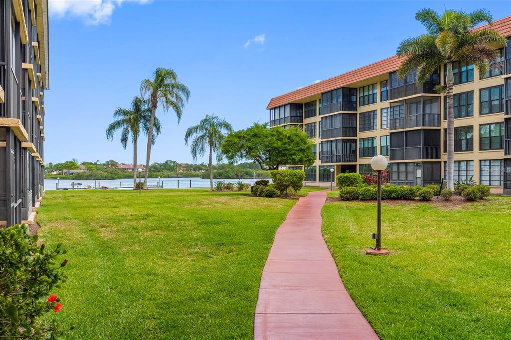 A paved walkway leads through a grassy courtyard between two multi-story apartment buildings near water — 19701 GULF BOULEVARD UNIT 307, INDIAN SHORES, FL
