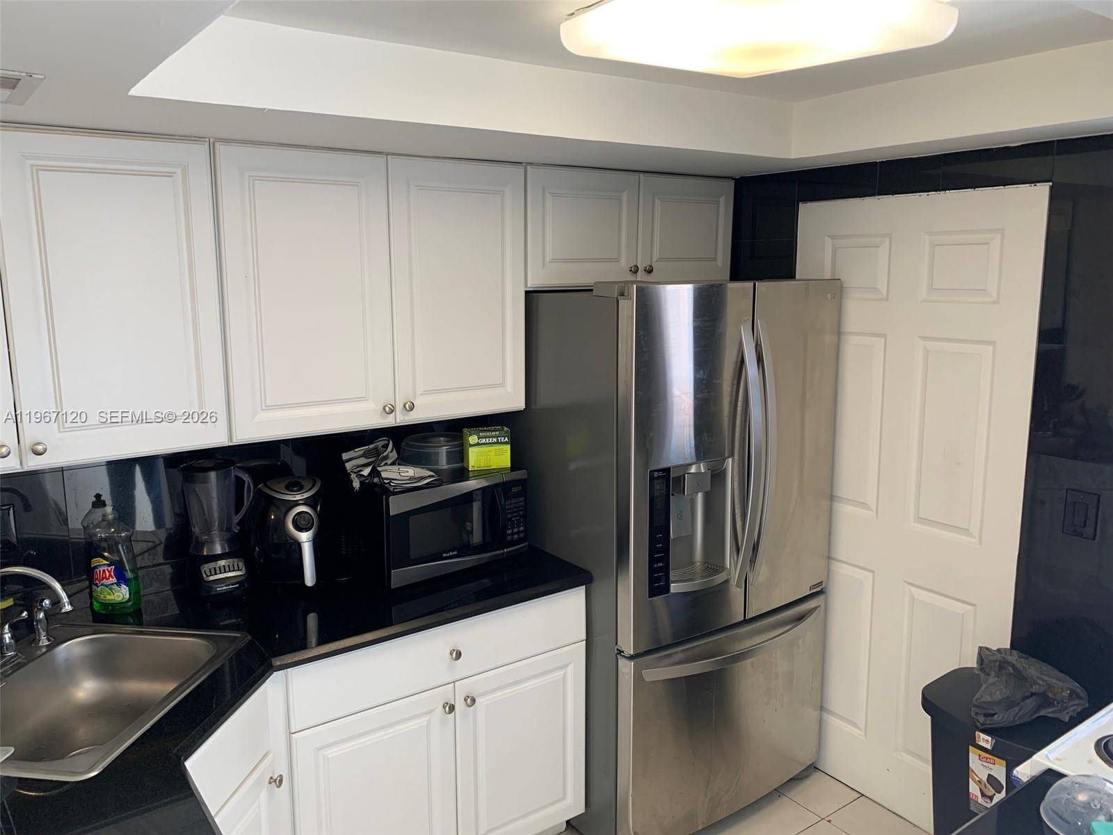 A kitchen features white cabinets, black countertops, a stainless steel refrigerator, and a sink — 1333 E Hallandale Blvd # 143, Hallandale Beach FL 33009, HALLANDALE BEACH, FL