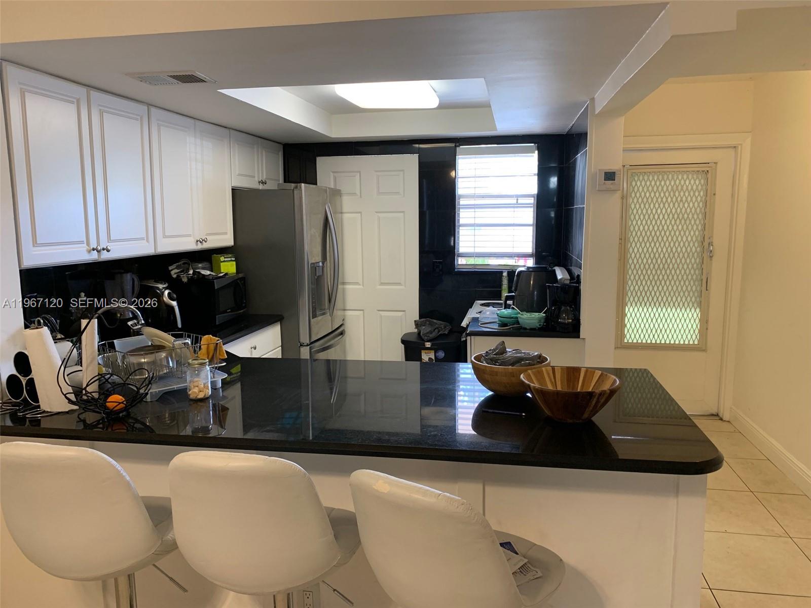 A kitchen features white cabinets, black countertops, a stainless steel refrigerator, and three bar stools — 1333 E Hallandale Blvd # 143, Hallandale Beach FL 33009, HALLANDALE BEACH, FL