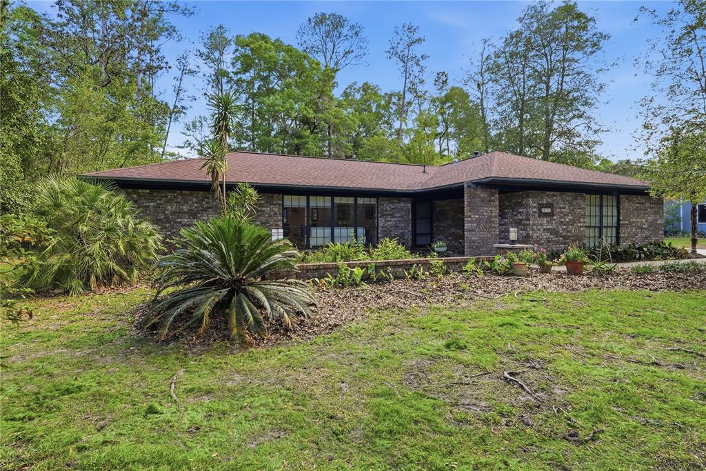 A single-story brick house with a dark shingled roof sits behind a landscaped front yard — 3705 NW 25TH AVENUE, GAINESVILLE, FL