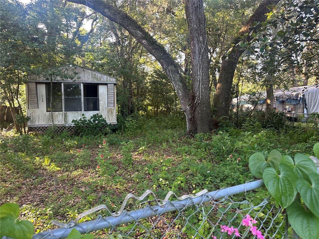 A weathered mobile home sits in a wooded, overgrown lot behind a chain-link fence — 1128 N JACKSON AVENUE S, SPRING HILL, FL
