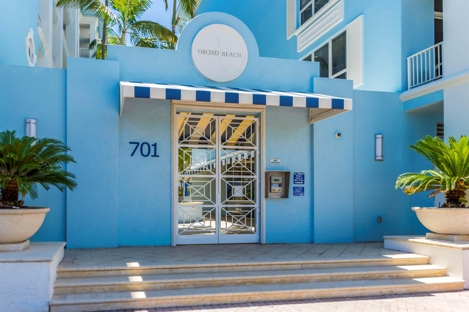 The entrance to a blue building features a striped awning, white metal doors, and potted plants — 701 SE 21st Avenue # 204, Deerfield Beach FL 33441, DEERFIELD BEACH, FL
