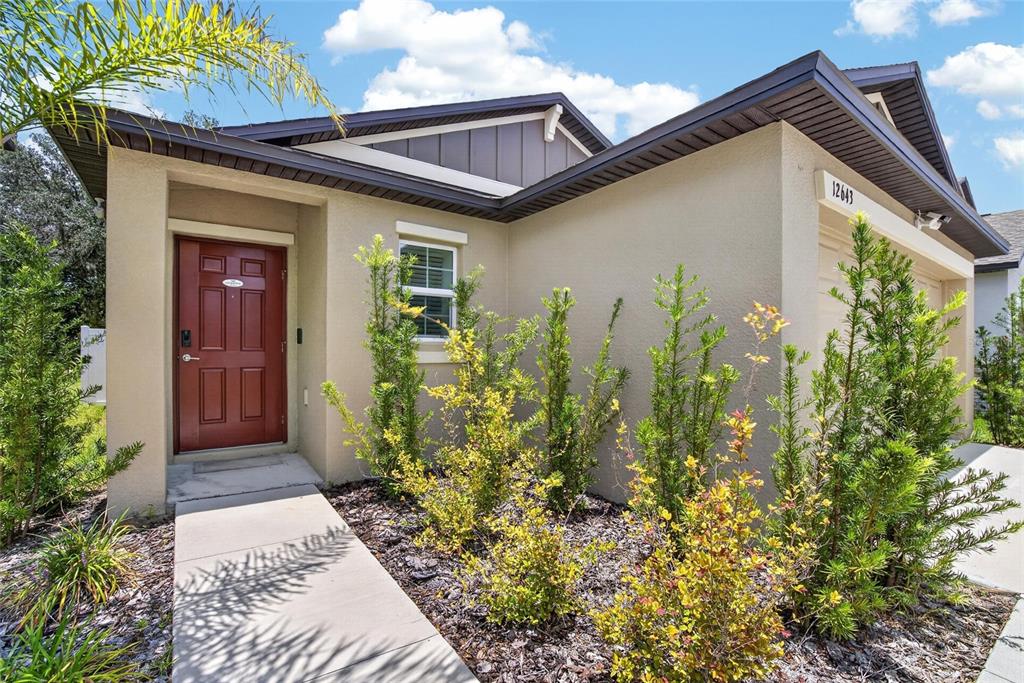 A tan single-story house features a red front door, a concrete walkway, and landscaping — 12643 MAPLE BONSAI DRIVE, RIVERVIEW, FL