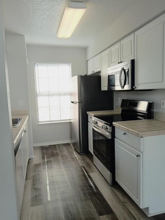 A galley kitchen features white cabinets, stainless steel appliances, tile countertops, and wood-look flooring — 2931 RIO GRANDE TRL UNIT D, KISSIMMEE, FL