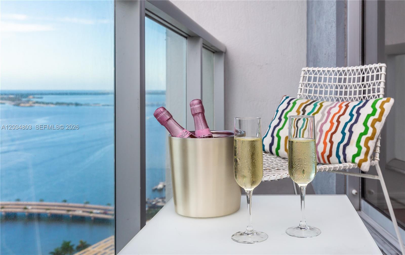 A balcony features a small table with champagne, two glasses, and a chair with a patterned pillow — 485 Brickell Ave # 3211, Miami FL 33131, MIAMI, FL