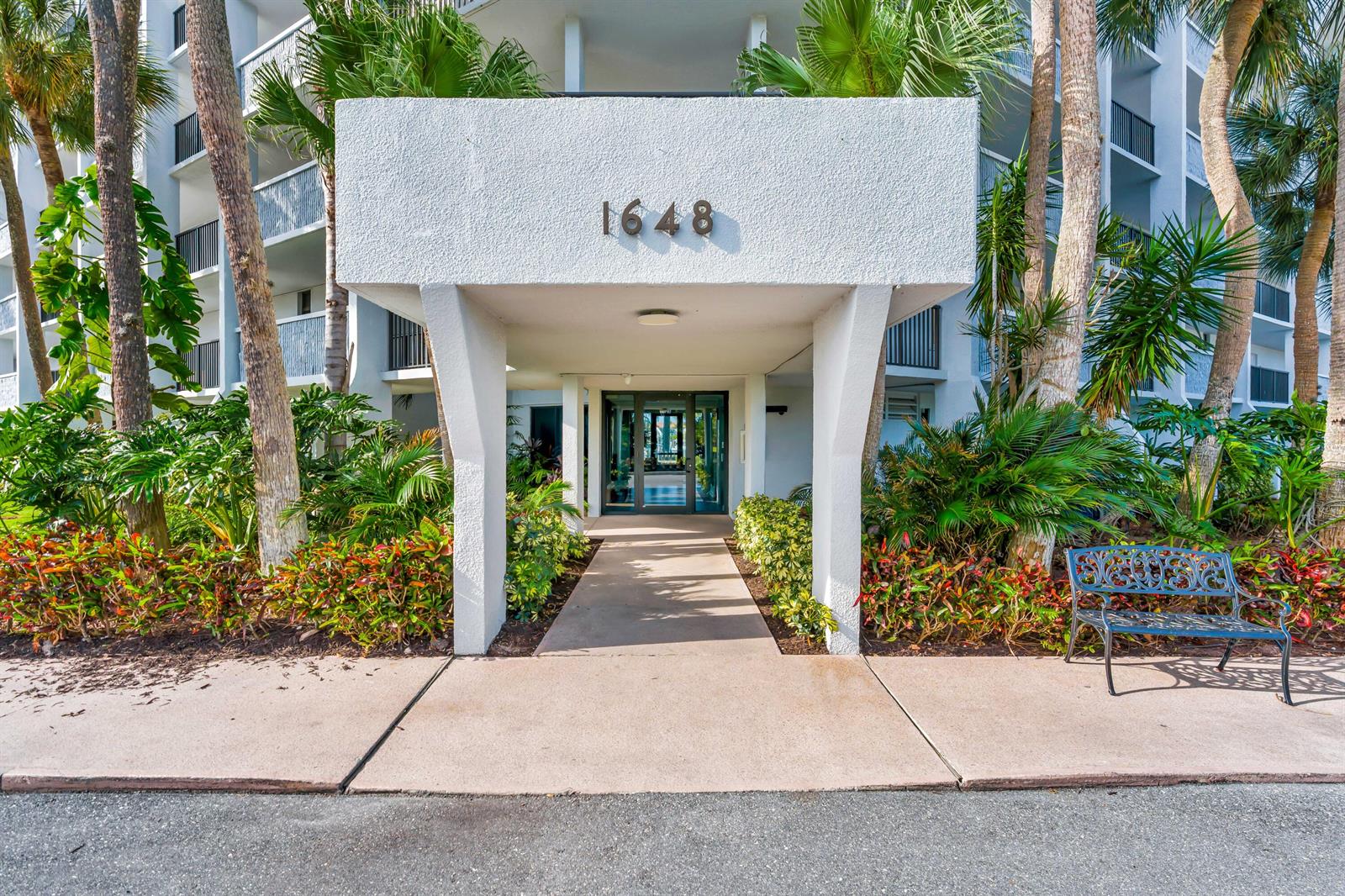 The exterior entrance of a multi-story building features a white concrete overhang and landscaping — 1648 Jupiter Cove Drive # 608b, Jupiter FL 33469, JUPITER, FL