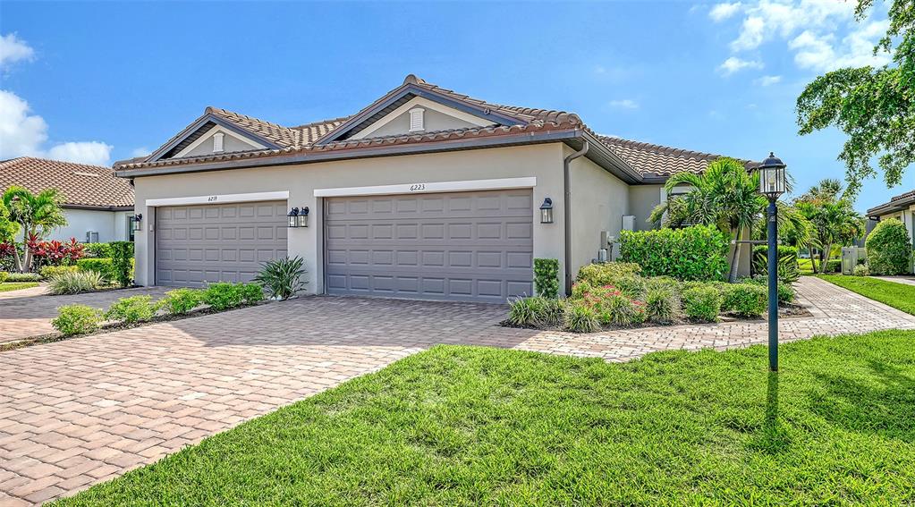 A beige duplex house features two garage doors and a paved driveway under a blue sky — 6223 PLATEAU COURT, BRADENTON, FL