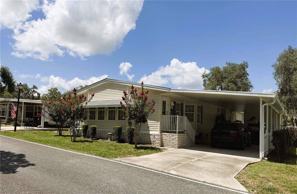 A single-story manufactured home with a carport, light siding, and blooming crepe myrtle trees — 103 ORCHID WAY, LEESBURG, FL