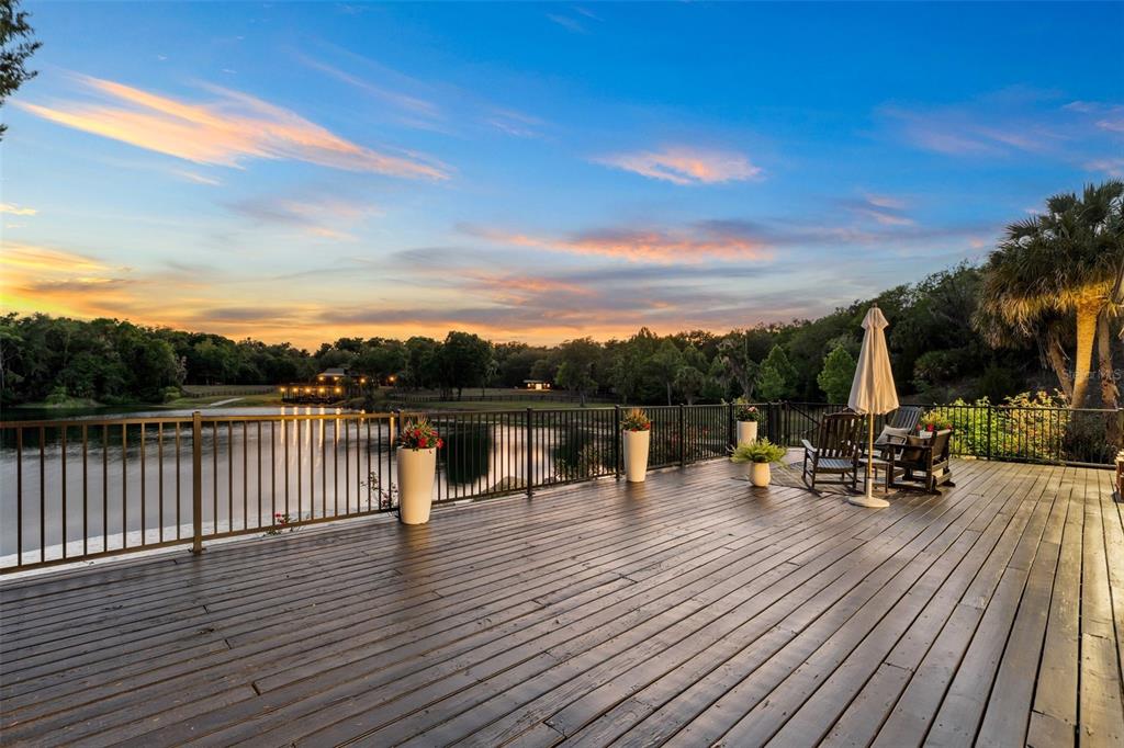 A wooden deck overlooks a lake at sunset with a metal railing and patio furniture — 17995 NE 24TH TERRACE, CITRA, FL