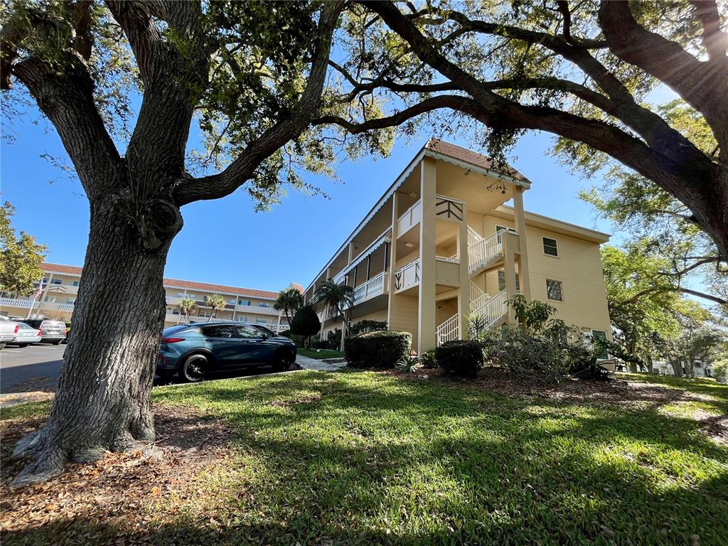 A multi-story apartment building with yellow siding and exterior staircases sits behind large oak trees — 2041 AUSTRALIA WAY W UNIT 49, CLEARWATER, FL