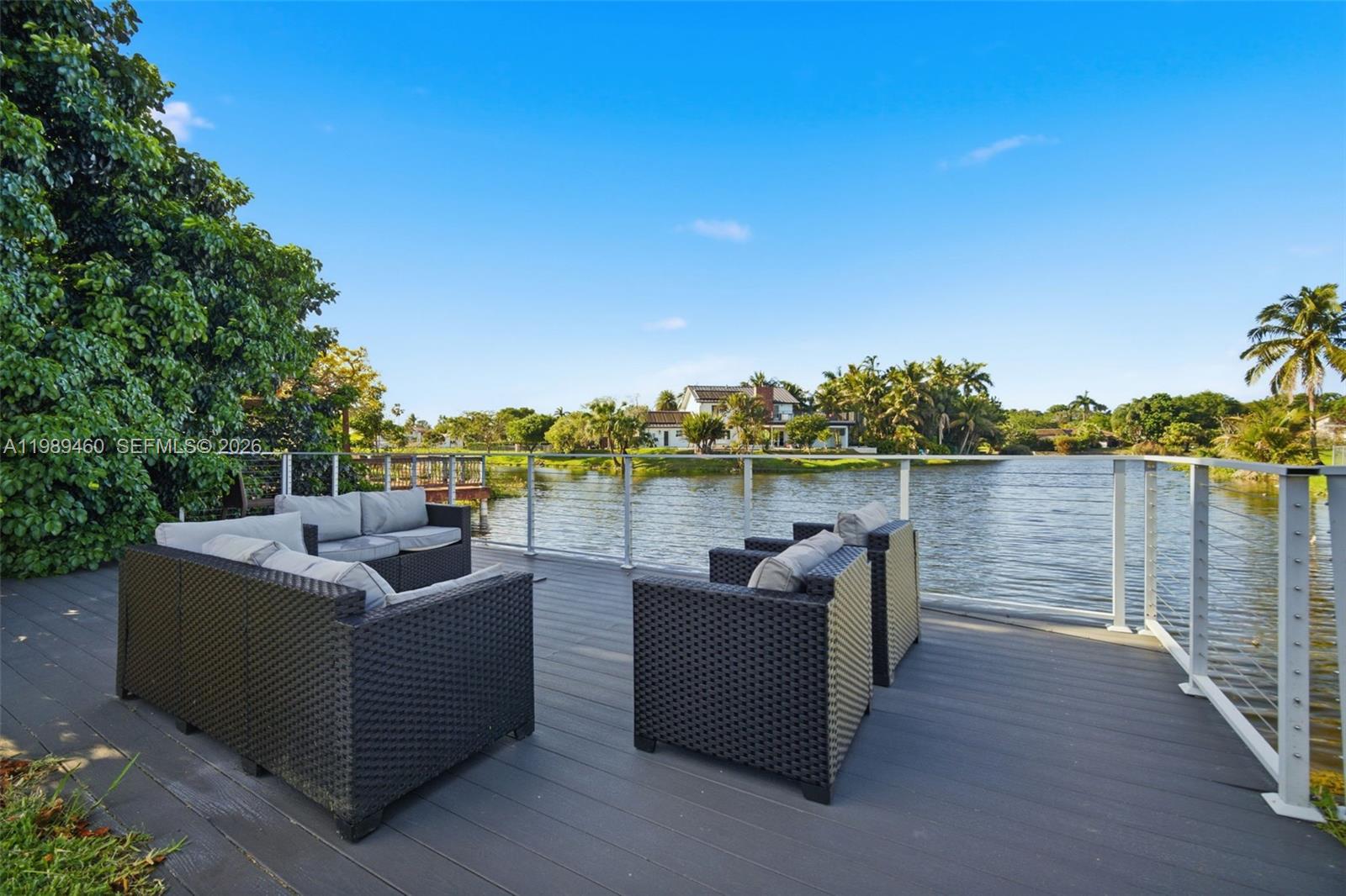 An outdoor deck with wicker furniture overlooks a lake with a house in the distance — 2201 SW 115th Ter, Davie FL 33325, DAVIE, FL