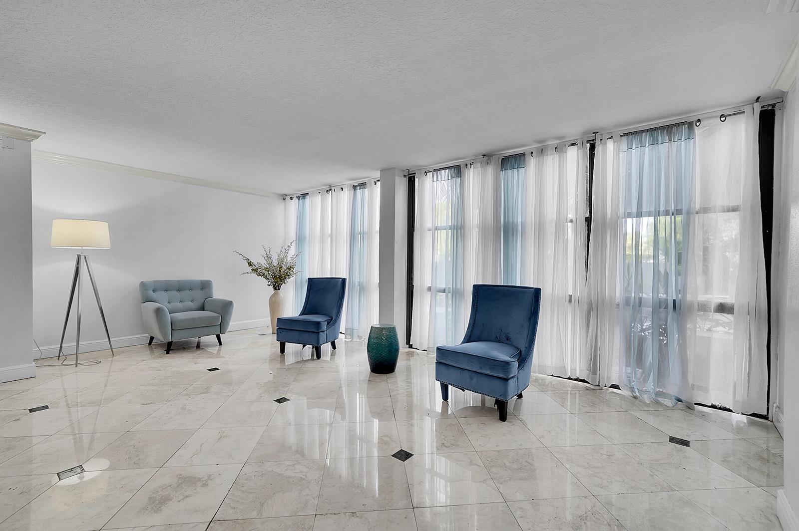 A bright living room features white marble tile floors, blue accent chairs, and floor-to-ceiling curtains — 1000 Parkview Drive # 306, Hallandale Beach FL 33009, HALLANDALE BEACH, FL