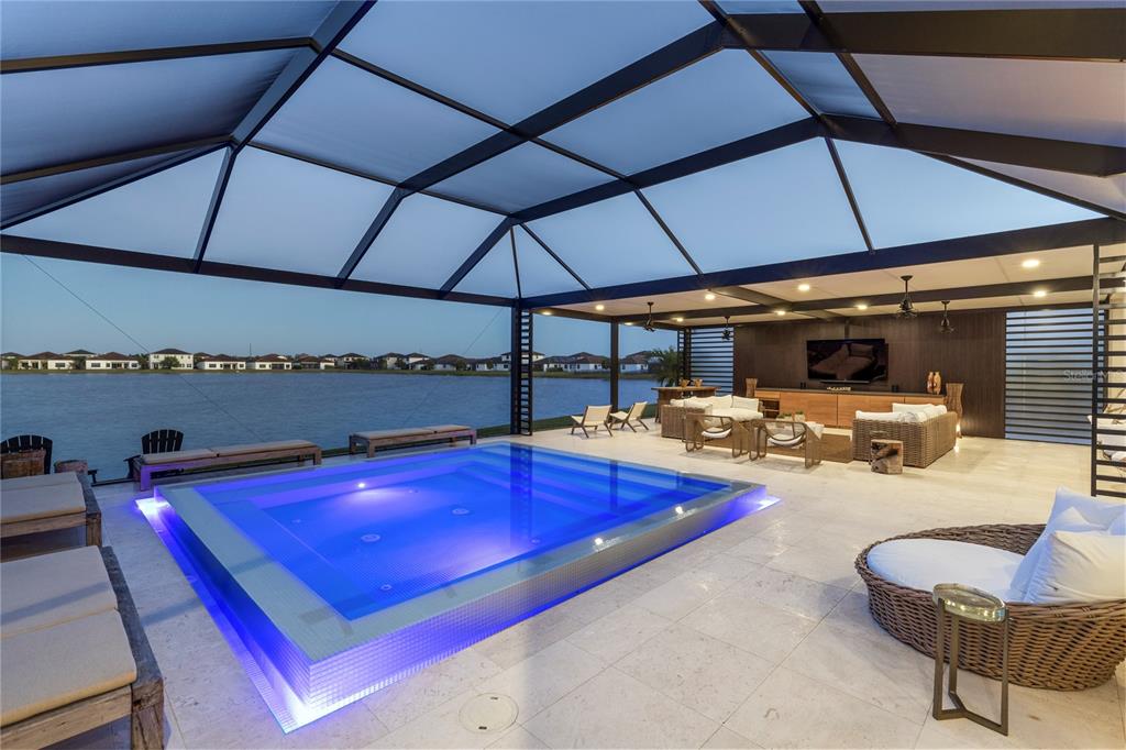 A covered patio features a rectangular pool with blue lighting overlooking a lake at dusk — 4796 SOPHIA DRIVE, AVE MARIA, FL