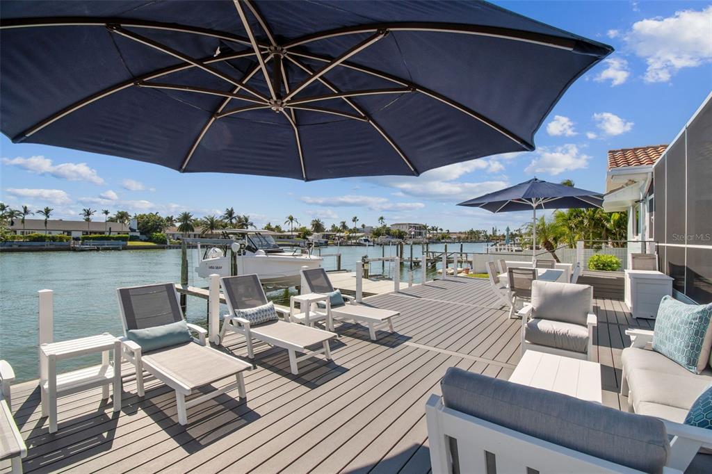 A wooden deck features lounge chairs, patio furniture, and umbrellas overlooking a canal with boats — 320 BELLE ISLE AVENUE, BELLEAIR BEACH, FL