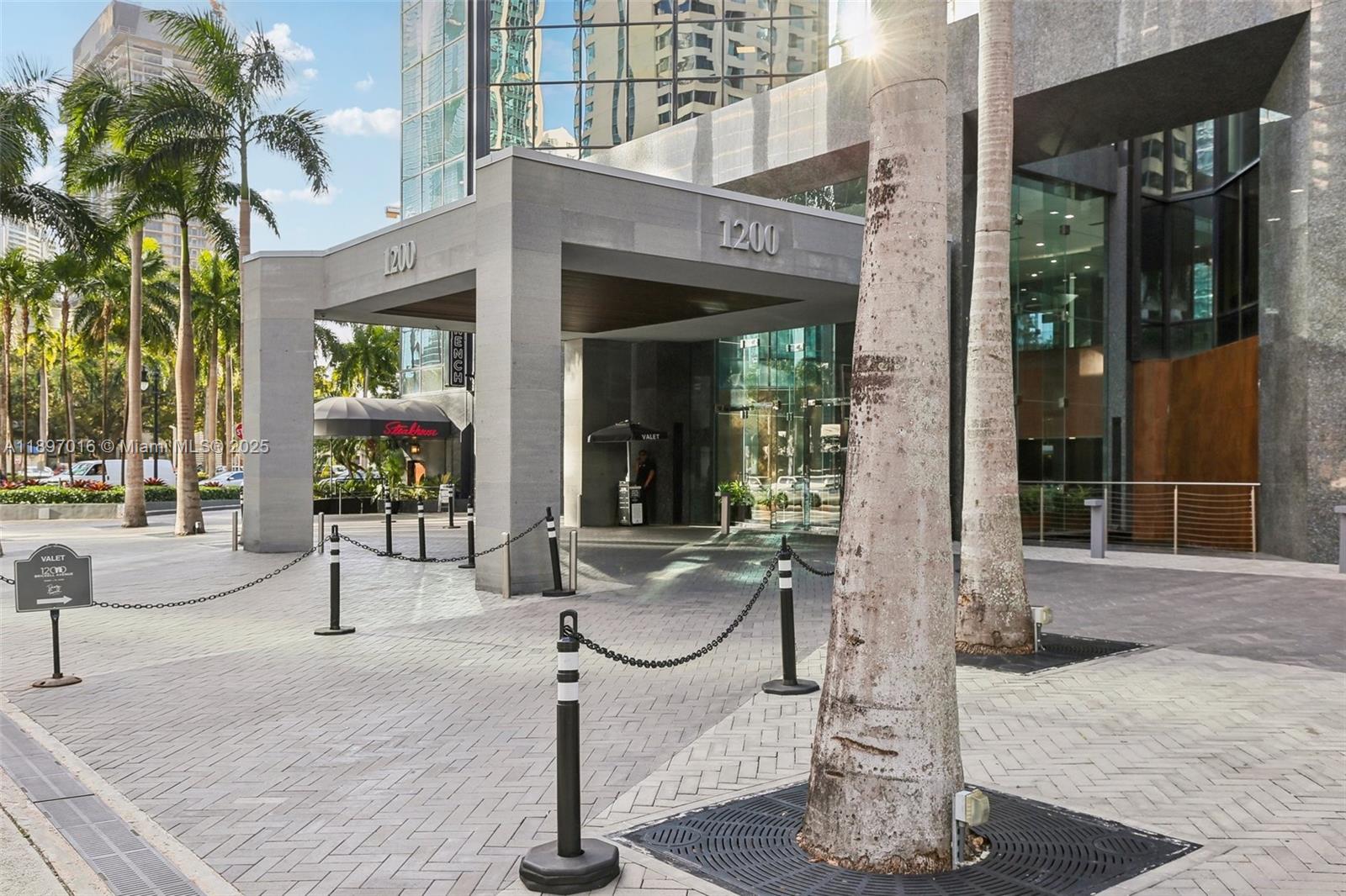 The exterior entrance of a modern building features a concrete porte-cochère and palm trees — 1200 Brickell Ave # D, Miami FL 33131, MIAMI, FL