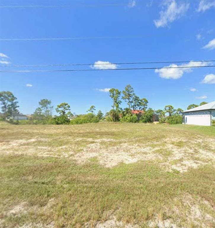 A vacant, grassy residential lot under a blue sky with scattered trees and nearby houses — 3112 NW 17TH PLACE, CAPE CORAL, FL