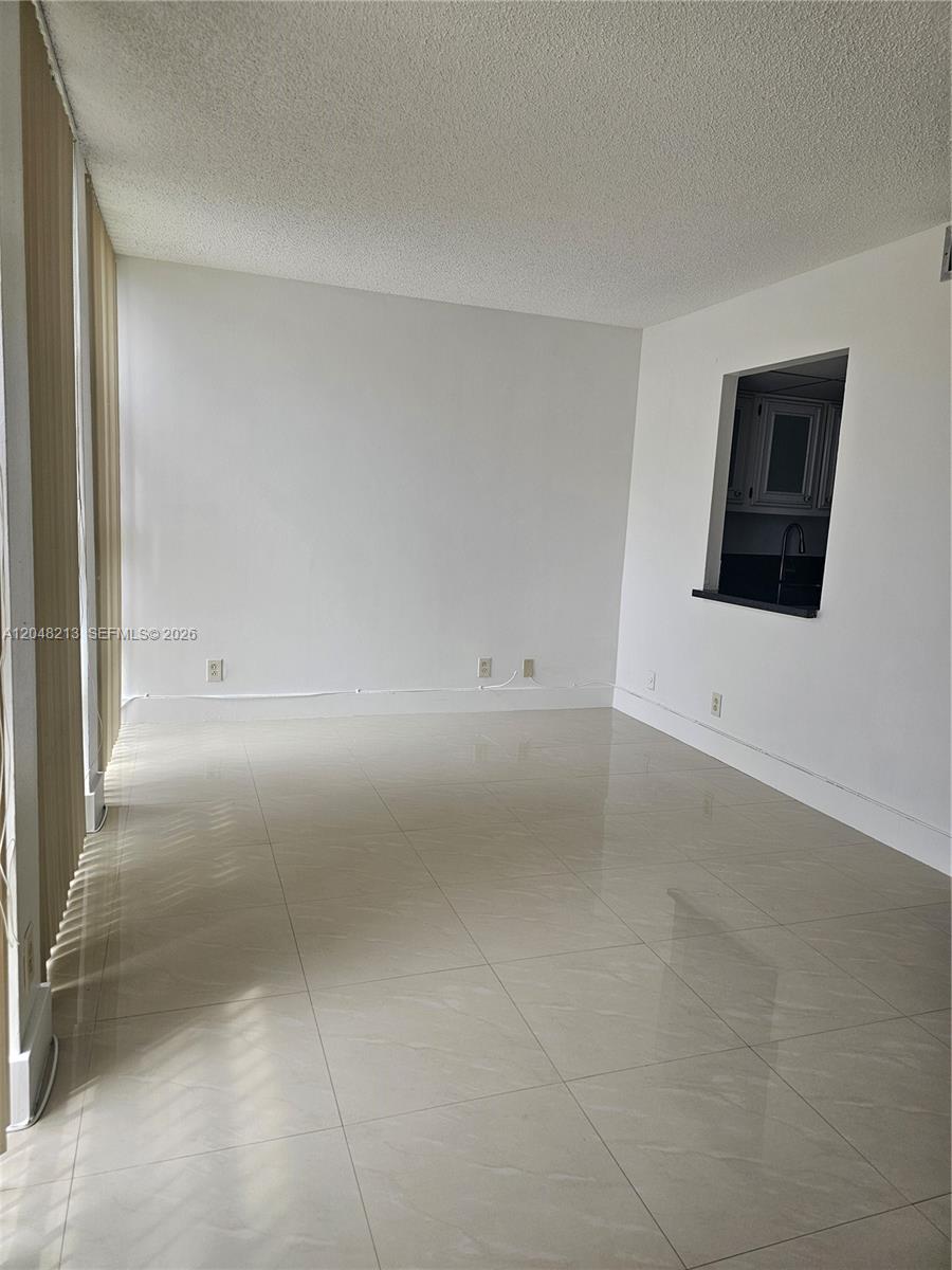 A bright, empty room features light-colored tile flooring, white walls, and a kitchen pass-through window — 801 Three Islands Blvd # 218, Hallandale Beach FL 33009, HALLANDALE BEACH, FL