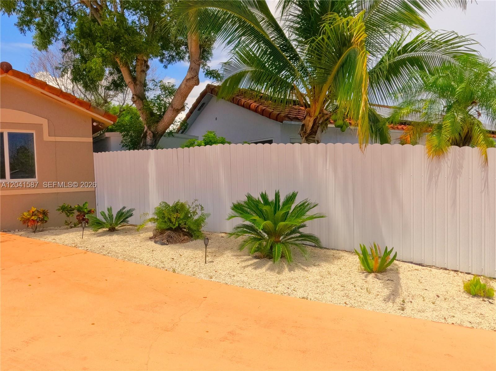 A white vinyl fence borders a gravel garden bed with various tropical plants and palms — 17700 SW 145th Ave, Miami FL 33177, MIAMI, FL