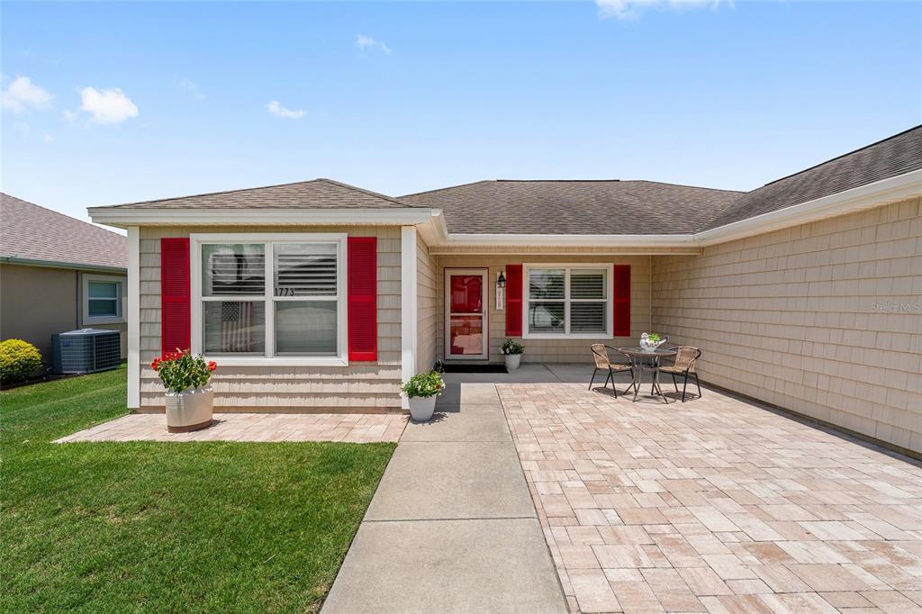 A single-story house exterior features tan shingle siding, red shutters, and a paved front patio — 1773 HOLLOW BRANCH WAY, THE VILLAGES, FL