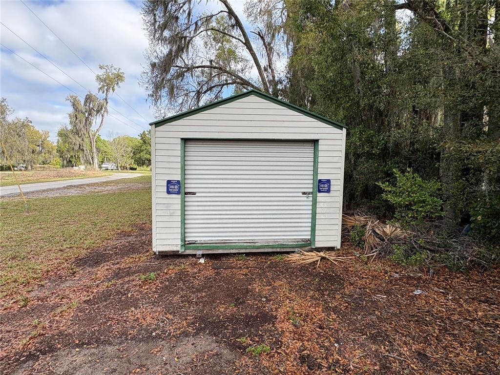 A beige storage shed with a roll-up door sits on a dirt lot near trees — 131 N C 470, LAKE PANASOFFKEE, FL