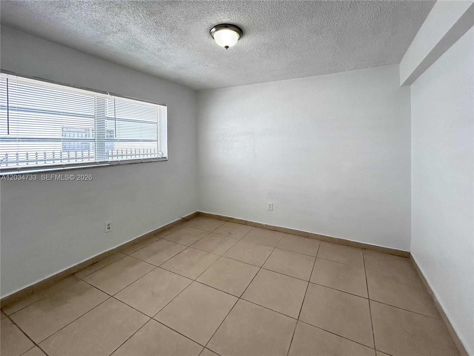 An empty room features white walls, beige tile flooring, and a window with blinds — 8287 NE Miami Ct # 102, Miami FL 33138, MIAMI, FL