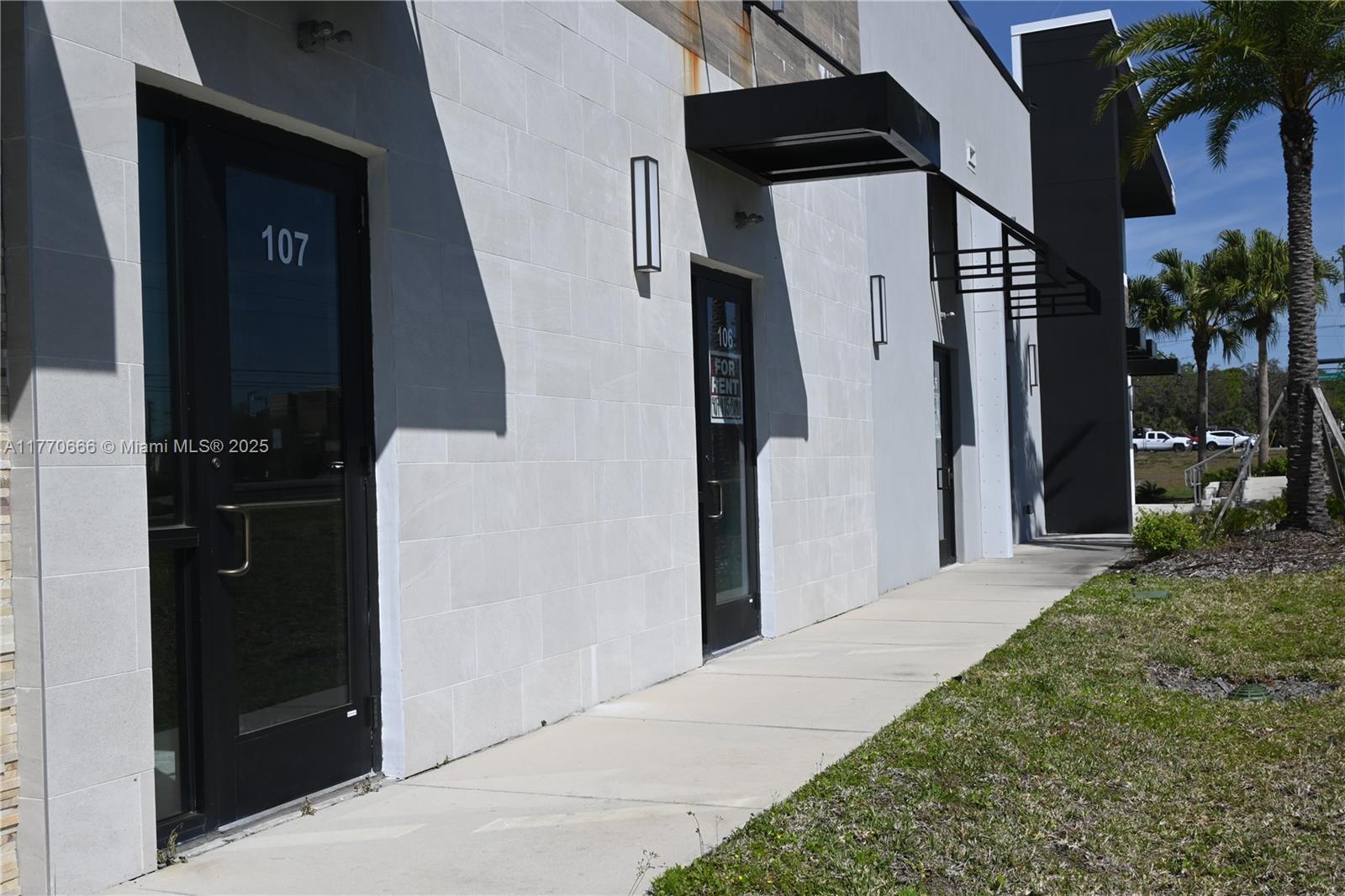 The exterior of a modern commercial building features glass doors, metal awnings, and palm trees — 3370 ROUSE RD # 107, Orlando FL 32819, ORLANDO, FL