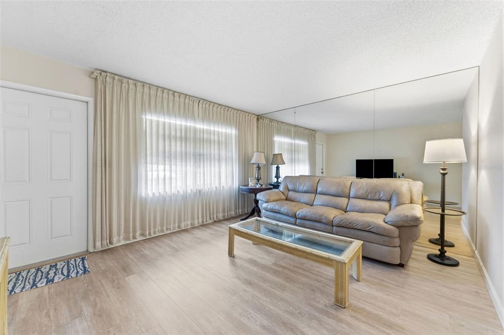 A living room features light wood-look flooring, a beige sofa, a glass coffee table, and mirrors — 24862 US HIGHWAY 19 N UNIT 806, CLEARWATER, FL