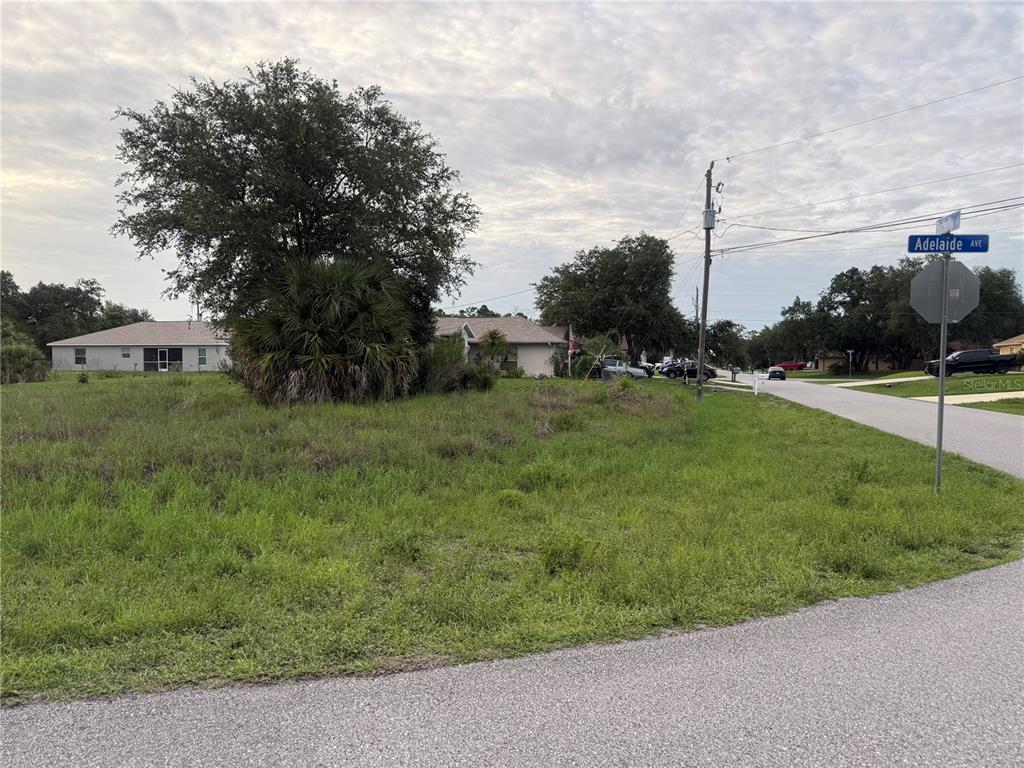 A vacant, grassy residential corner lot features a large tree and a street sign — ADELAIDE AVENUE, NORTH PORT, FL