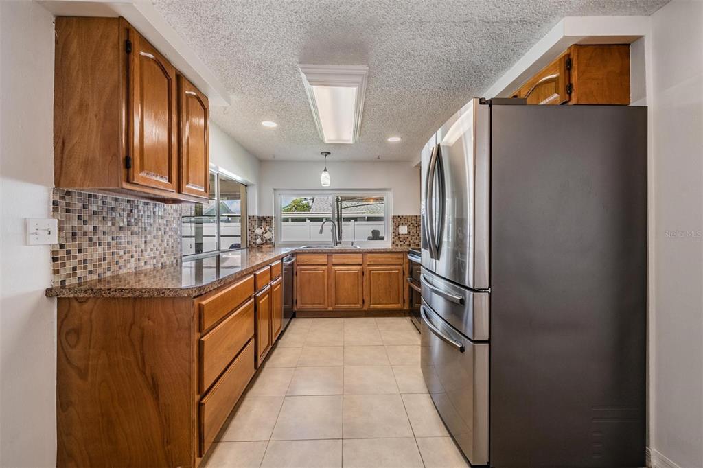 A galley kitchen features wood cabinets, granite countertops, a mosaic backsplash, and stainless steel appliances — 3849 CLAREMONT DRIVE, NEW PORT RICHEY, FL