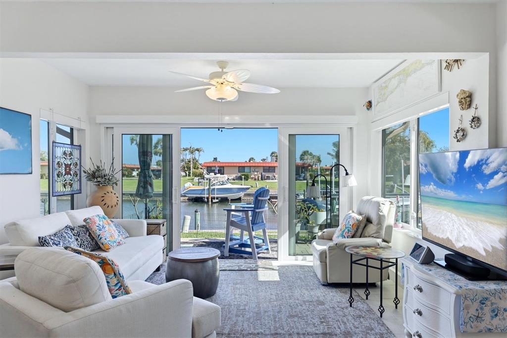 A living room features white furniture, a ceiling fan, and sliding doors overlooking a canal — 9528 ANTIQUA COVE UNIT 9528, BRADENTON, FL
